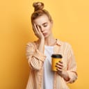 A tired girl in white t-shirt and orange striped shirt holding a cup of beverage resting her face on her right hand