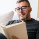 A middle aged man with glasses and a bear sitting on sofa reading a book