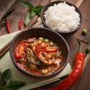 A meat soup with chilly with a bowl of rice placed on a wooden table with peppers as garnish