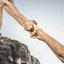 close-up of a person offering assistance by extending a hand to help another individual climb a mountain.