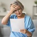 shocked older woman reacting to news in a letter, touching her head in despair while sitting on a couch