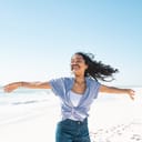 A young, smiling woman standing on the beach with her arms open wide