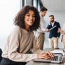 Woman smiling into the camera while working on laptop with her coworkers in the background