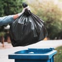 person putting a black garbage bag into a blue trash bin