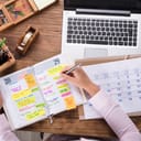 Close-up Of Businesswoman Writing Schedule In Calendar Diary On Desk