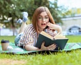 Surprised woman reads book while lying on a blanket in the park