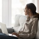 young woman with eyeglasses sitting on a couch with her computer, working remotely and gazing outside the window