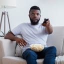 surprised young man sitting on a couch with a remote in hand and popcorn on his lap