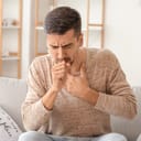 A young man sitting on a sofa holding his chest and coughing