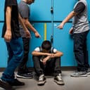 A sad high school boy sitting on the floor in front of lockers as his peers point fingers at him