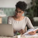 Woman looking at laptop while sitting at table with a notebook and pen in her hands