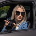 A blonde woman sitting in a car holding a camera