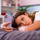Depressed teenage girl lying on her bed at home and scrolling through social media on her mobile phone