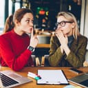 Brunette hipster blogger told secret to wondered colleague and showing sign shh during collaborating at laptop devices in coworking