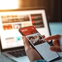 Person holding their phone in front of a laptop cross-referencing a new article.
