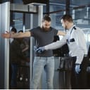 A security agent at an airport conducts a pat-down on a bearded male passenger after he passes through the metal detector scanner in the departures hall.