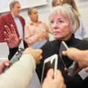 A mature businesswoman addressing reporters equipped with microphones and smartphones at a press conference.