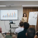 A confident female professional presenting a sales graph on a screen to an attentive office audience, conveying success and teamwork.