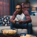 A young man playing a video game while sitting on a sofa with a bowl of chips in front of him