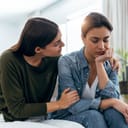young woman comforting her sad female friend on the sofa at home