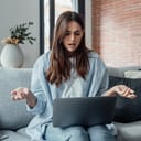 An annoyed young woman sitting on a couch in her living room, visibly frustrated while working on her laptop.