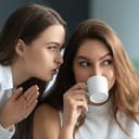 A woman whispering into another woman's ear while she is drinking a cup of coffee