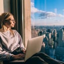 Female digital nomad with a laptop on a skyscraper window sill, enjoying the cityscape