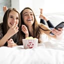 Two girls laying in bed and eating popcorn while watching a movie
