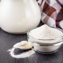 Powdered milk in a small glass bowl next to a jug of milk and a spoon full of milk powder