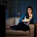 young lovely girl holding popcorn box and looking at tv screen enjoying movie showing puzzled emoticon sitting in living room comfortable sofa eating snack at night.