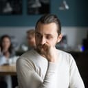 A worried man sitting alone in an office while his coworkers are sitting together in the background