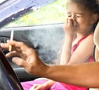 Small girl pinching her nose with eyes closed as she sits inside a car with a driver who is smoking