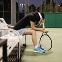 A sad, young female tennis player sitting on a bench at the side of the court leaning her head on her racket