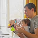 father attempting to work from home with his baby daughter in his arms, and the other one in a chair next to them