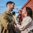 Smiling young couple flirting and having fun at a rooftop party with friends, drinking beer, enjoying and having a pleasant conversation. Handsome guy greeting his joyful girlfriend at celebration