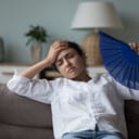 A young woman sitting on a couch feeling hot trying to cool herself with a fan