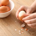 a woman peeling a boiled egg on a wooden table next to a small ceramic bowl with more boiled eggs