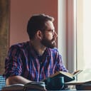 Bearded man holding a book and thoughtfully looking out the window
