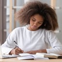 A young girl writing in a notebook with books and a laptop opened on the desk in front of her