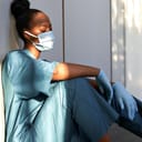 A tired, exhausted African scrub nurse, wearing a face mask, blue uniform, and gloves, sits on the hospital floor.