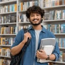 Portrait of cheerful male international Indian student with backpack, learning accessories standing near bookshelves at university library or book store during break between lessons. Education concept