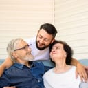 A man hugging his parents who are sitting on a couch