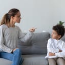 A strict mother scolds her mischievous young son as they sit on a sofa at home.