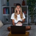 A young woman sitting on her bed, with a confused face expression as she watches a movie on her laptop.