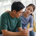 sad young man dealing sitting on a couch in his living room and a woman providing support, holding his hand and lightly hugging him