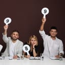 A panel of judges holding different score signs at a table, set against a brown background.