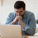 A worried man sitting in front of his laptop looking at the screen with his arms resting on the mouth