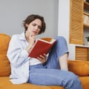 Puzzled pensive confused young woman 20s in white casual clothes sit on couch reading book put hand prop up on chin spending time in living room at home. Rest relax good mood leisure lifestyle concept