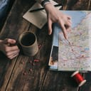 close-up of a woman's hands, planning a travel route on a map with pins and red string while holding a cup of tea on a wooden table
