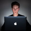 Man sitting at a Macbook computer in a dark room.